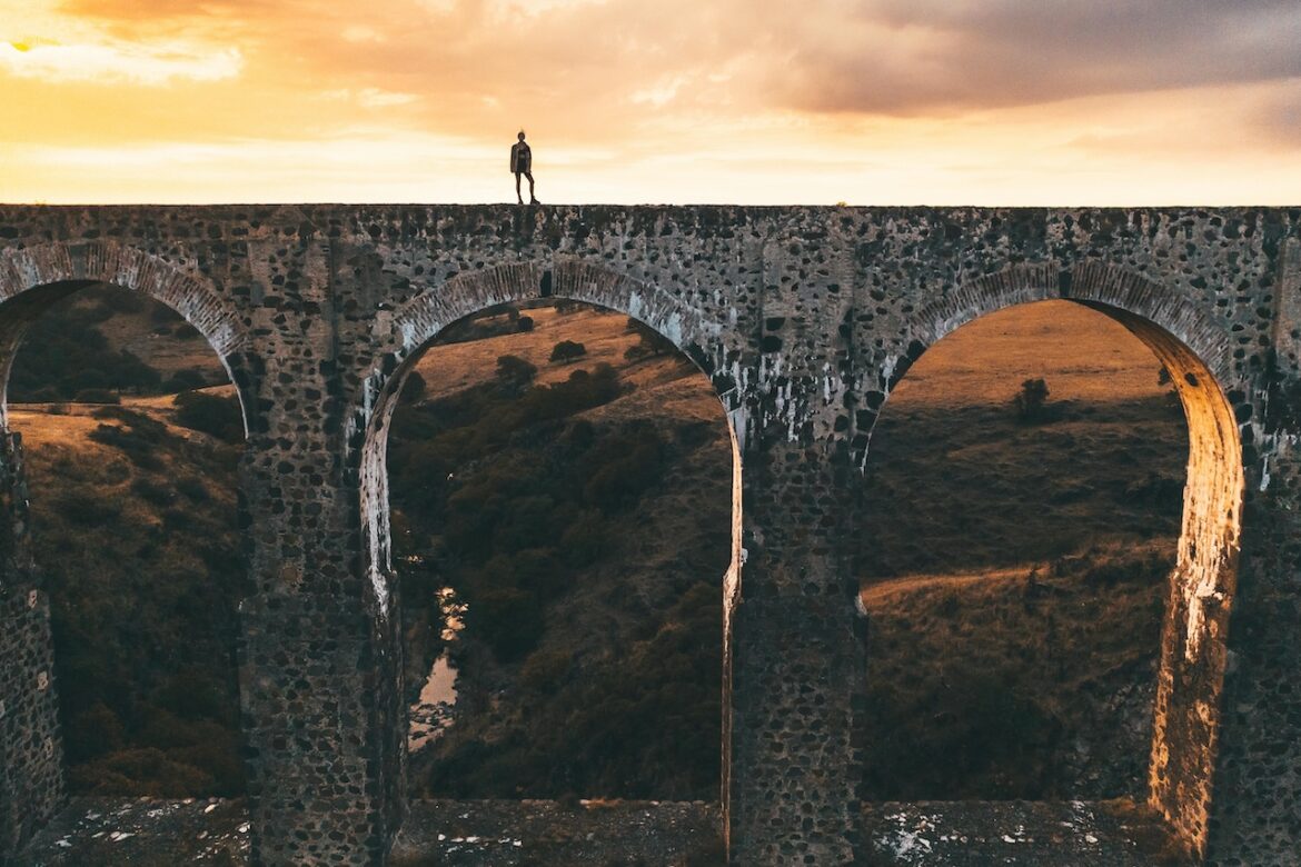Woman traveling on top of an aqueduct - a large stone structure which is broken up with various portals that allow for glimpses out toward a beautiful landscape. The aqueduct is meant to symbolize the ease which honoring your boundaries can bring to your life without closing you off to the outside world, relationships and opportunities.